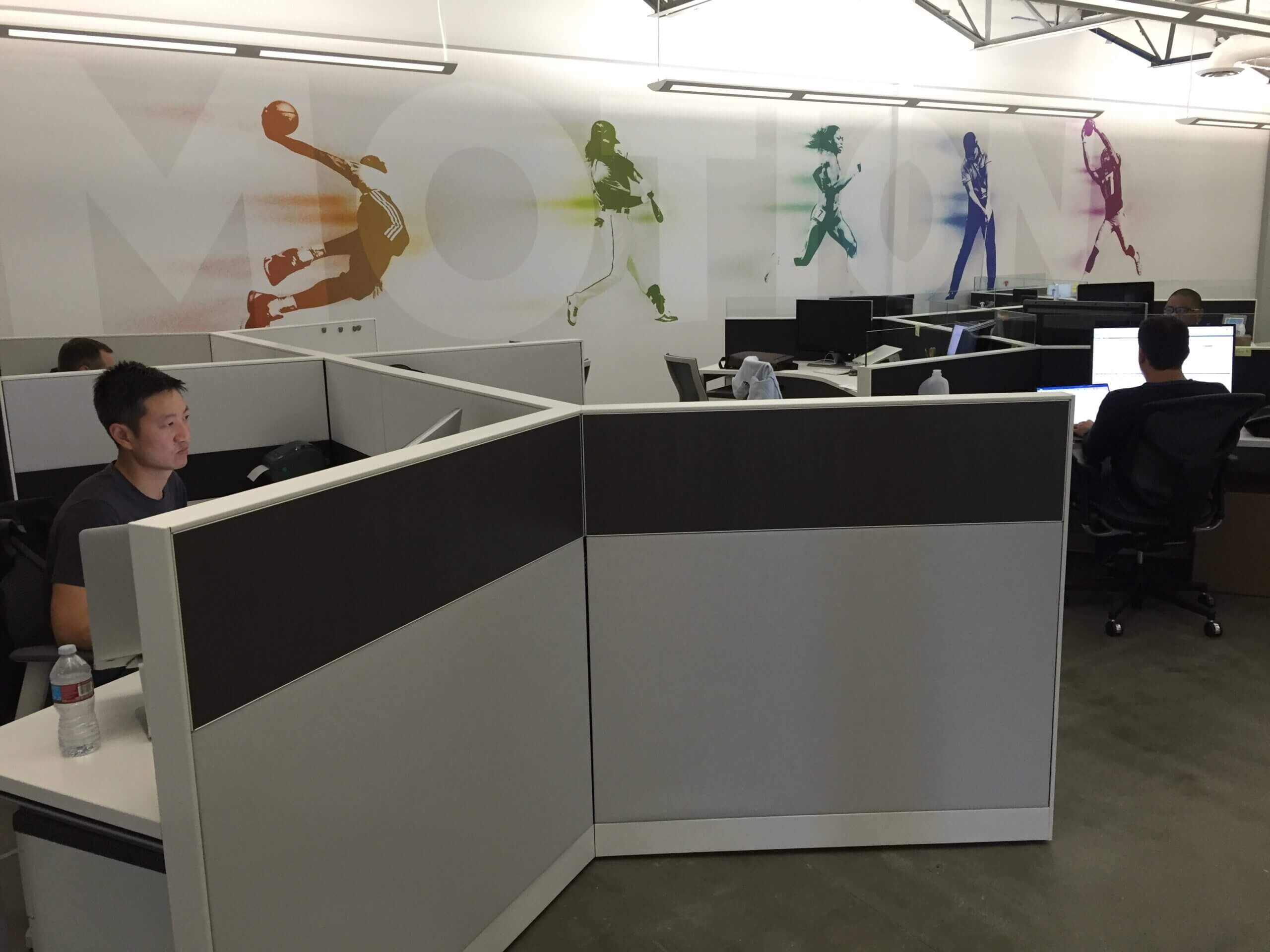 Employees sit at computers in their respective cubicles. The wall behind them displays colorful renditions of athletes in action.