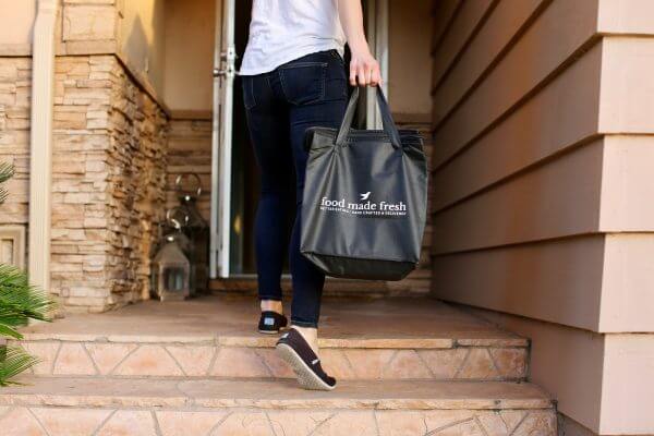 A delivery person carries a black Food Made Fresh bag to someone's front door