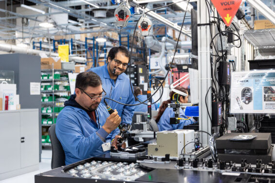 Two men in blue coats use an electronic tool on an assembly line