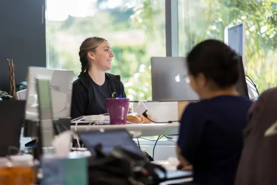 A worker sits at her desk working on her computer by a large window