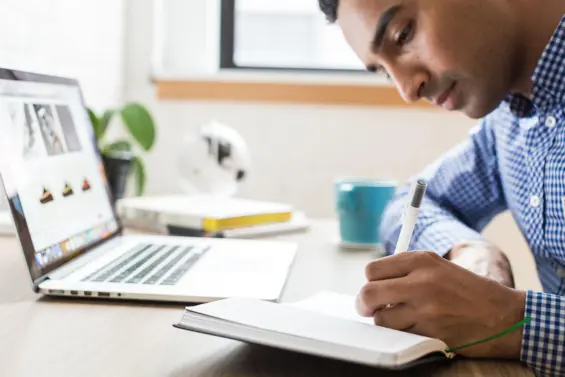 A man takes notes in a notebook while working on his laptop